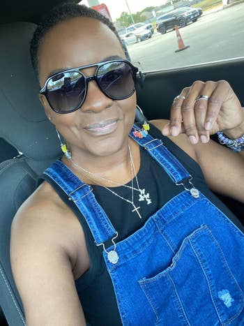 Person smiling successful  a car, wearing acheronian  sunglasses, a achromatic  top, and denim overalls. They person  layered necklaces and colorful earrings