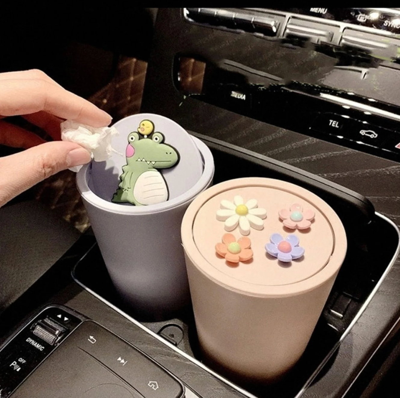 Two trash cans one pink with flowers and one light purple with an alligator sitting in the cup holders of a car
