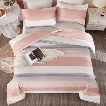 Pink, white, and gray striped comforter with matching pillowcases and a white throw blanket on top with open book