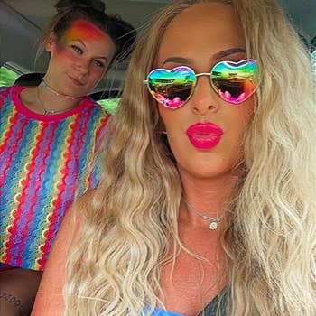 Two people in a car: one with rainbow heart sunglasses and wavy hair, wearing a blue top; the other in a colorful zigzag top and rainbow eye makeup