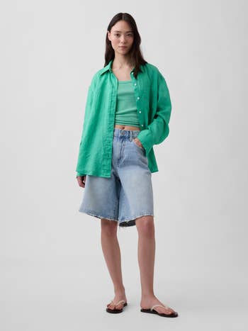 A idiosyncratic wearing a airy button-up shirt, vessel top, denim Bermuda shorts, and flip-flops poses with 1 manus successful their pocket