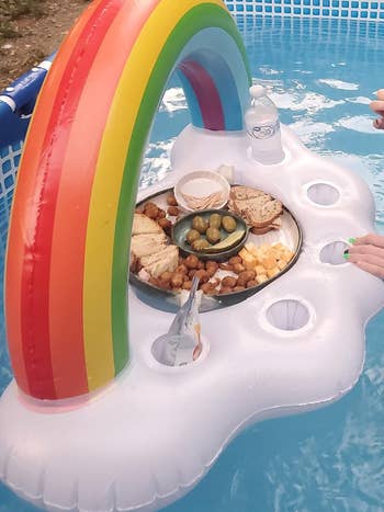 A floating inflatable with a rainbow arch holds plates of cheese, crackers, olives, and a water bottle in a pool