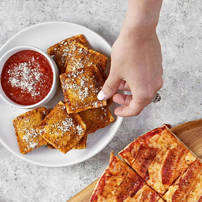 A manus reaches for a crispy ravioli adjacent to a vessel of dipping sauce, with a sliced pizza adjacent connected a woody tray
