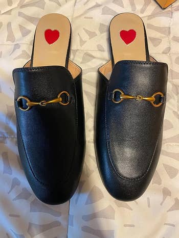 Close-up view of a pair of black leather mule loafers with gold horsebit detailing and red heart insoles