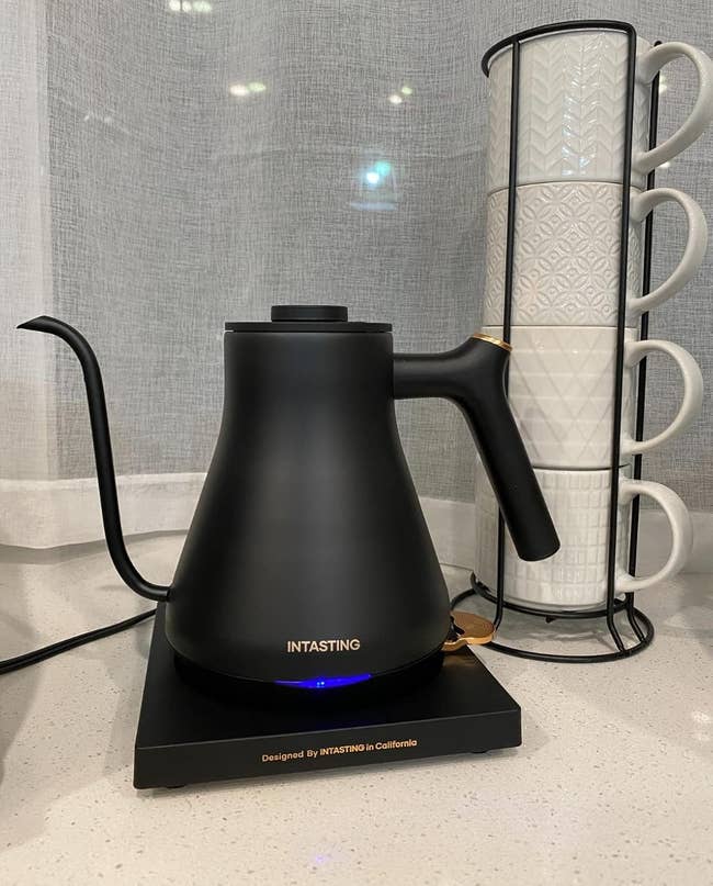 Electric kettle with a sleek plan connected a countertop adjacent to stacked ceramic mugs, suitable for modern kitchens
