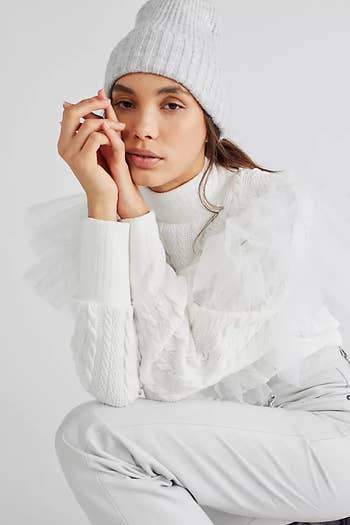 front of a model crouching while wearing a white knit turtleneck with tulle detailing and white jeans and a white hat