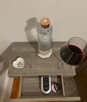 Reviewer image of product with slim drawer open and a remote and vaseline inside and a dish, water bottle, and glass of wine on top