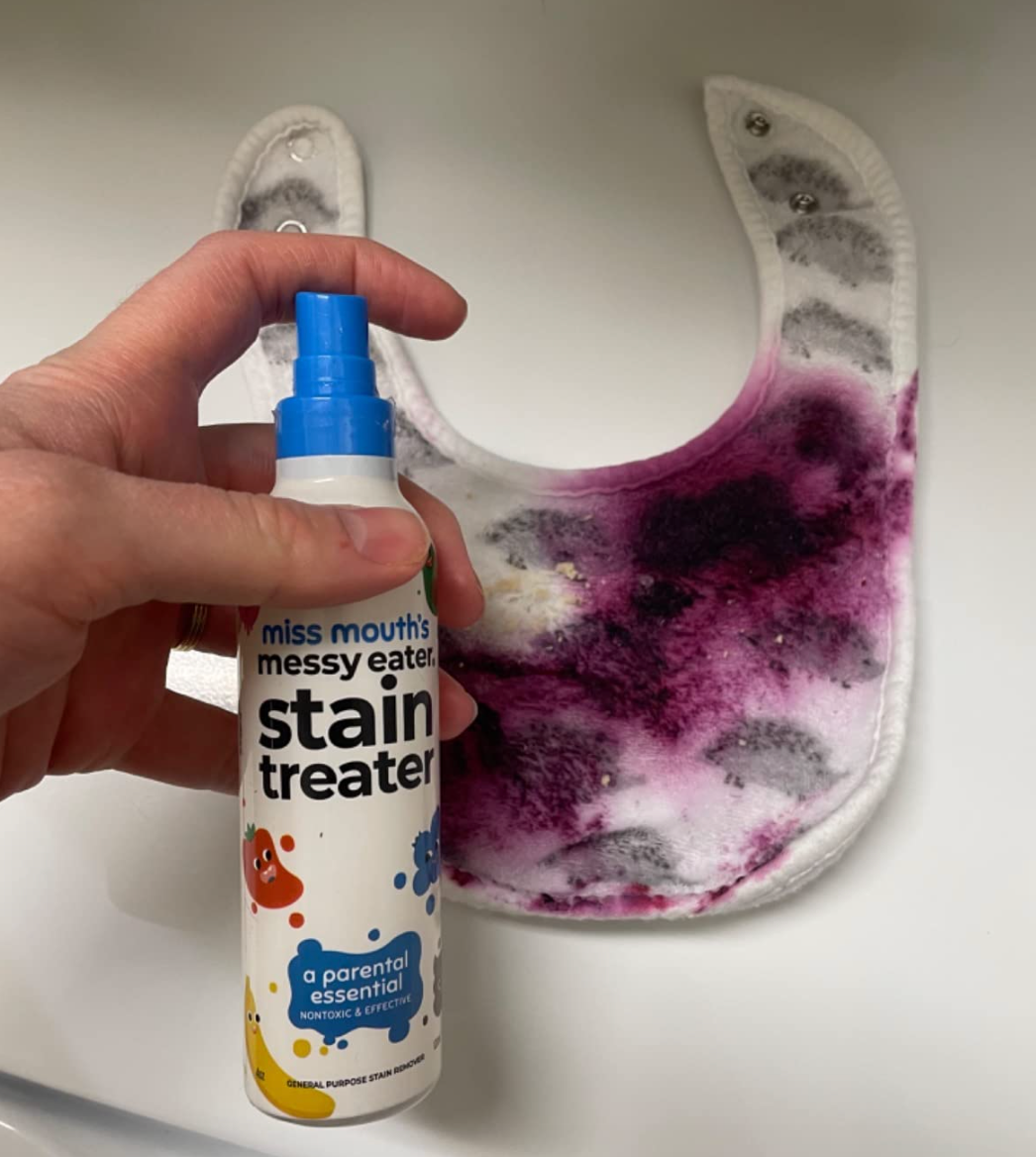 A hand holding a bottle of Miss Mouth's Messy Eater stain treater, with a stained bib in the background