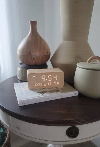 Wooden digital clock displaying 9:54 AM, 6/25 SAT 71°F on a round table with a book titled 