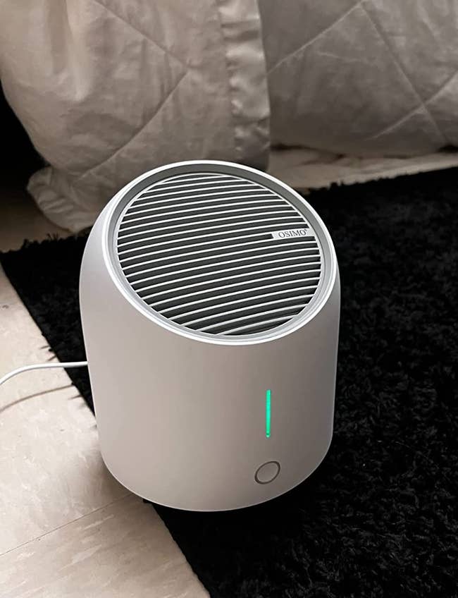 Reviewer image of white air purifier with green light on top of black carpet in front of bed with white blanket