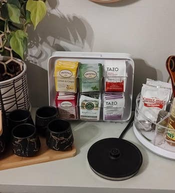 Assorted teas and kitchen items on a counter next to a potted plant, above cabinet drawers