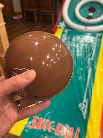 A hand holding a brown Skee-Ball with a Skee-Ball game setup in the background