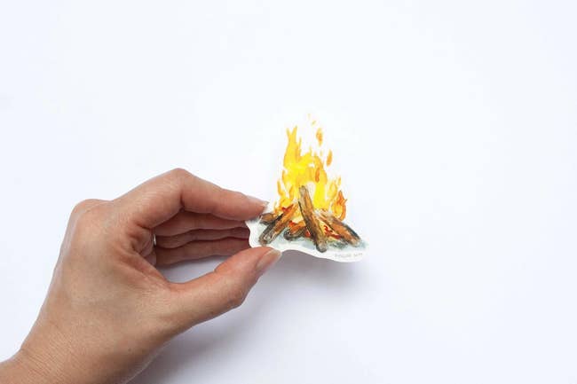 A hand holds a small sticker of a campfire, evoking a cozy, outdoor aesthetic