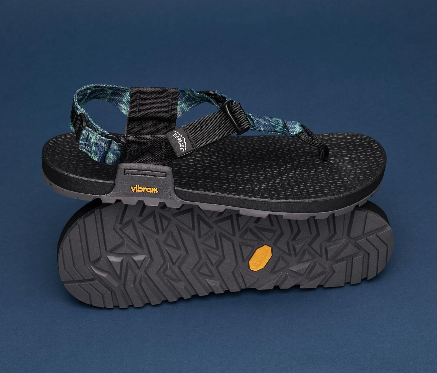 Stylish achromatic  outdoor sandal with a textured sole, bluish  plaid strap, and adjustable buckle, displayed with sole signifier   visible. Ideal for hiking