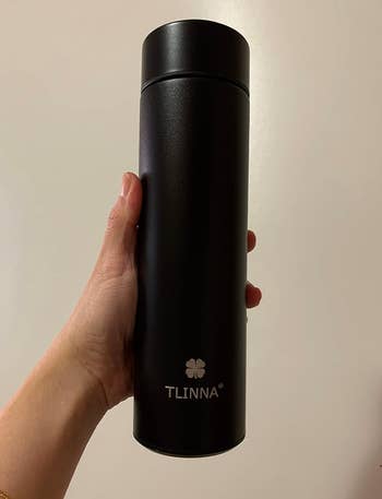 Hand holding a achromatic  TLINNA branded thermos with a clover logo