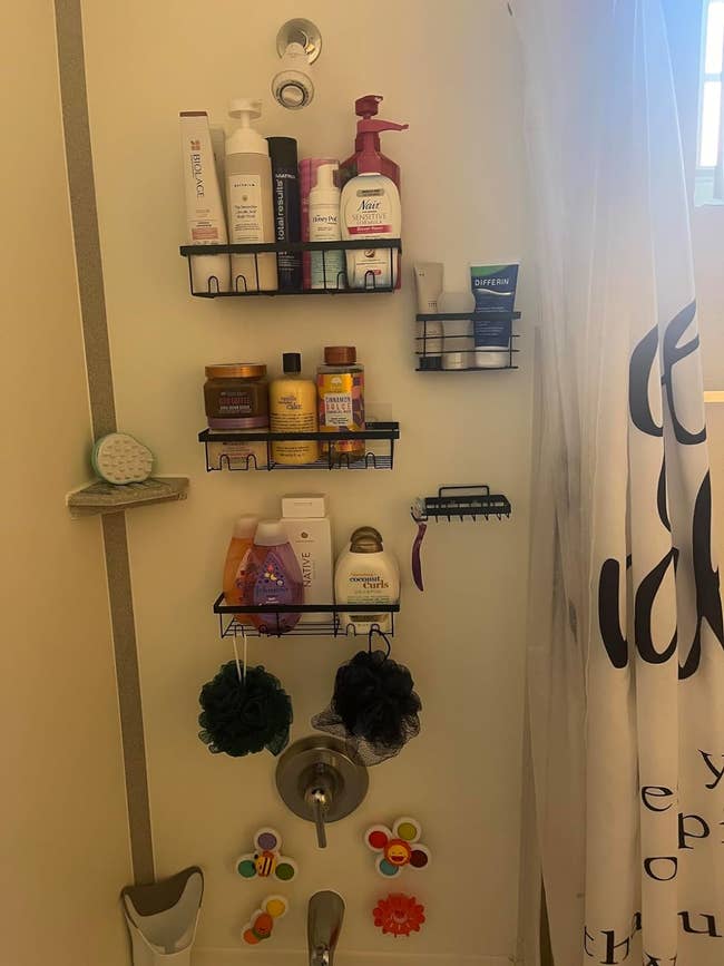 a reviewer's shower wall with five black adhesive shelves with different shampoos and bottles and two luffas hanging from the shelf