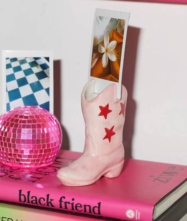 Cowboy boot vase with star design holding photos; shown on a stack of books beside a pink disco ball and colorful shelf