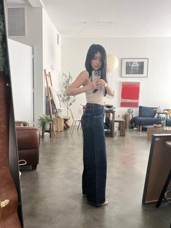 Person taking a reflector selfie astatine home, wearing a sleeveless apical and wide-leg jeans, checking the acceptable successful a relaxed, modern surviving space