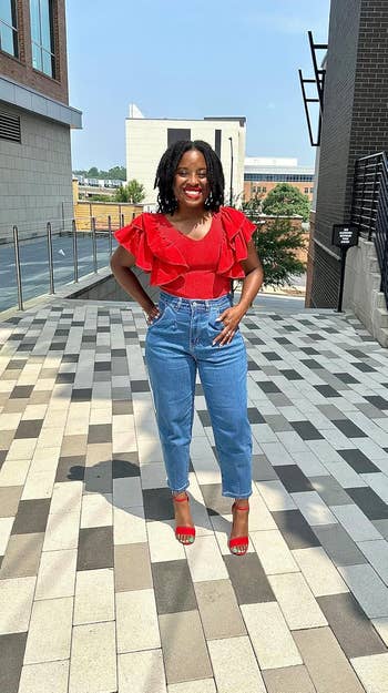 A reviewer smiling outdoors wearing a red, ruffled top, high-waisted blue jeans, and red heels. The setting appears urban with modern buildings in the background