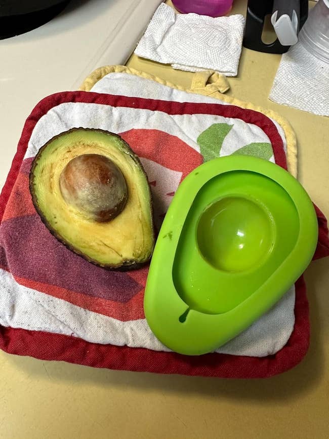 A halved avocado with the pit in one half, next to a green avocado saver on a kitchen towel