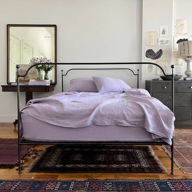 A well-made bed with iron frame, fresh purple linens, beside artwork and a floor mirror