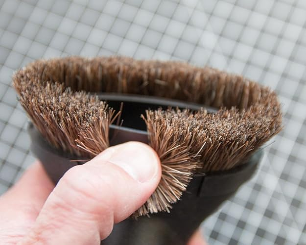 Hand holding a vacuum brushwood  attachment with dense bristles, against a checkered tile background. Ideal for cleaning and dusting surfaces