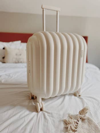 A beige ribbed hard-shell suitcase with extended handle, placed upright on a bed, suggesting travel preparation