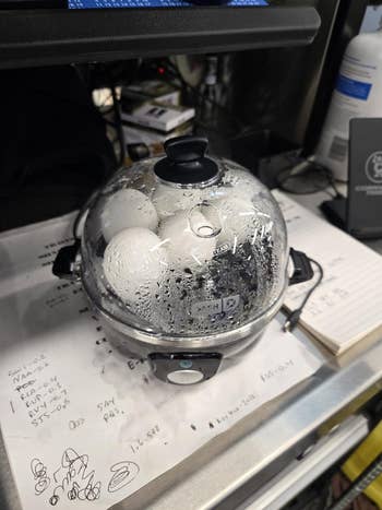 Electric ovum  cooker with eggs steaming inside, placed connected  a countertop with papers and notes successful  the background