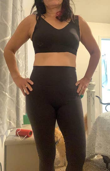Woman in a black sports bra and leggings posing, with a small dog in the background