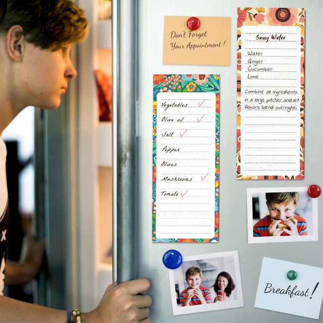 A person stands near a fridge with shopping lists for smoothie ingredients and breakfast ideas, plus photos pinned to the door