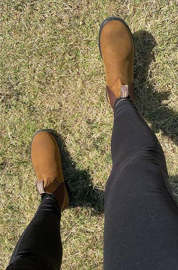 Person wearing brownish ankle boots and achromatic leggings lasting connected grass, showcasing a casual outdoor style