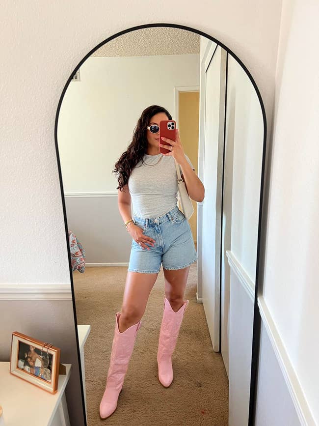 Person taking a reflector  selfie wearing a achromatic  sleeveless top, denim shorts, and pinkish  knee-high boots, showcasing a casual outfit style