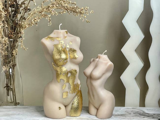 two nude Venus candles one shorter than the other and the taller one with gold flakes on it