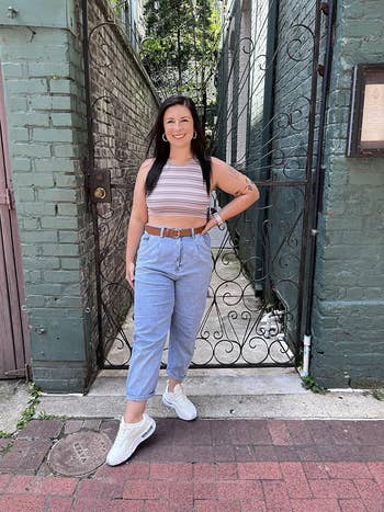 A reviewer is standing outside against a brick wall in casual attire, wearing a sleeveless top, belted jeans, and sneakers, with one hand on their hip