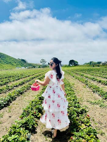 Person in a floral dress holding a basket at a strawberry field, posing as they walk between rows of plants