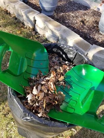 Green plastic leaf scoops filled with dry leaves being emptied into a black trash bag for outdoor cleaning