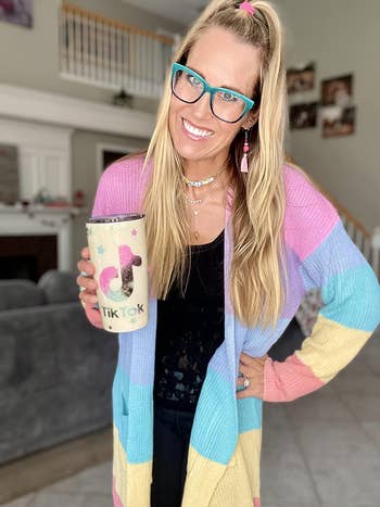 A reviewer wearing the rainbow cardigan with one of their hands on their waist and the other holding a cup