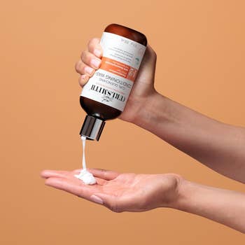 A person's hand dispenses creamy product from a Clearly Innovative Dominodic Wash bottle onto their palm against a plain background