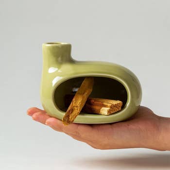 Hand holding a modern, creaseless ceramic incense holder with incense sticks inside. The plan is sleek and minimalistic, perfect for location decor