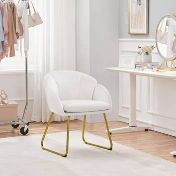 A modern home office setup featuring a white cushioned chair with gold legs, a white desk with decor items, and a clothing rack with various garments in the background