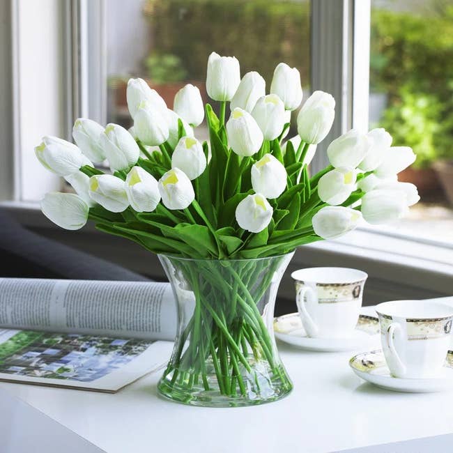 Vase with white tulips on a table, accompanied by a magazine and two teacups with saucers, near a window