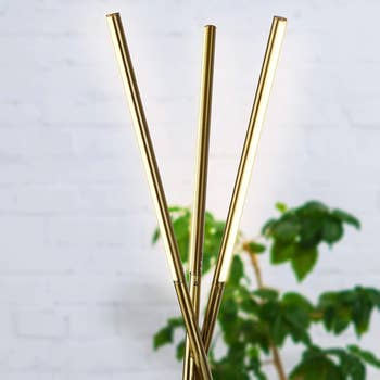 Modern LED floor lamp with three slim, vertical gold rods in front of a blurred plant background