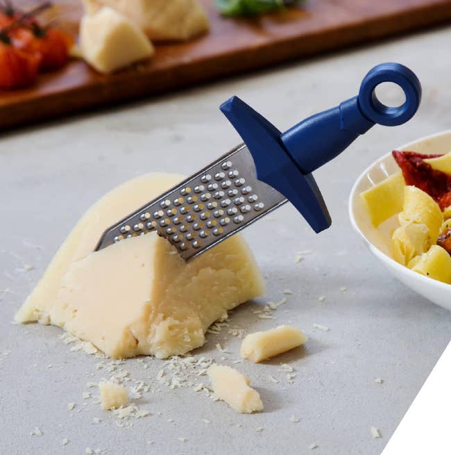 Cheese wedge on a counter with a blue-handled grater. A bowl of mixed ingredients is nearby, suggesting kitchen prep or a recipe inspiration