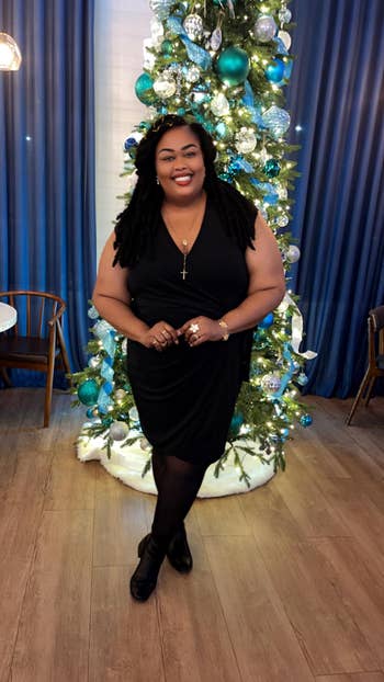 Person in elegant black dress poses in front of a decorated Christmas tree, smiling warmly