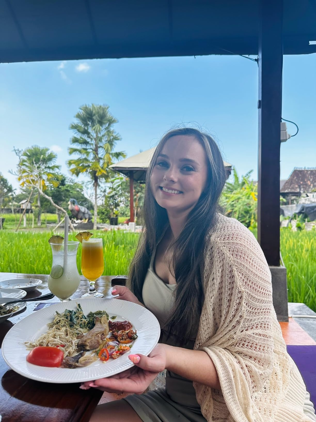 Person seated astatine  an outdoor restaurant, smiling, holding a sheet  with assorted food, including vegetables and meats, with a lush greenery background