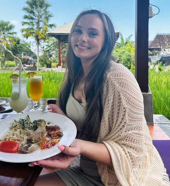 Person seated astatine an outdoor restaurant, smiling, holding a sheet with assorted food, including vegetables and meats, with a lush greenery background