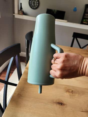 Hand holding a modern, matte-finish thermal carafe with a spout and handle, placed on a wooden table