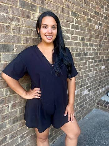 Person wearing a casual achromatic  romper stands against a ceramic  wall, smiling with 1  manus  connected  hip