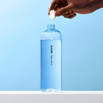 Hand dropping a cleaning tablet into a Blueland glass cleaner bottle filled with water against a plain background