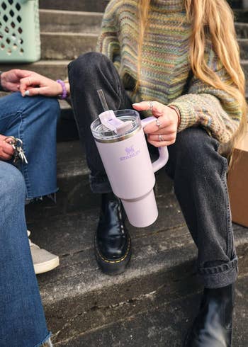 Person sitting on steps holding a purple stanley mug with a straw, wearing a striped sweater and black boots